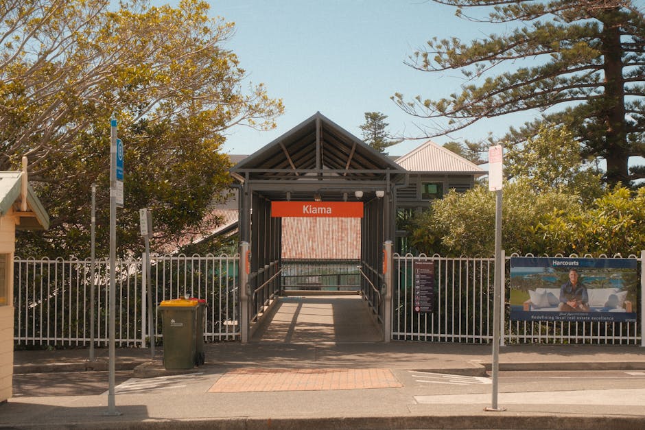 A straight pedestrian walkway with dark asphalt surface and metal handrails on both sides, leading over a railway bridge towards a train station platform in the background. The bridge's sides consist