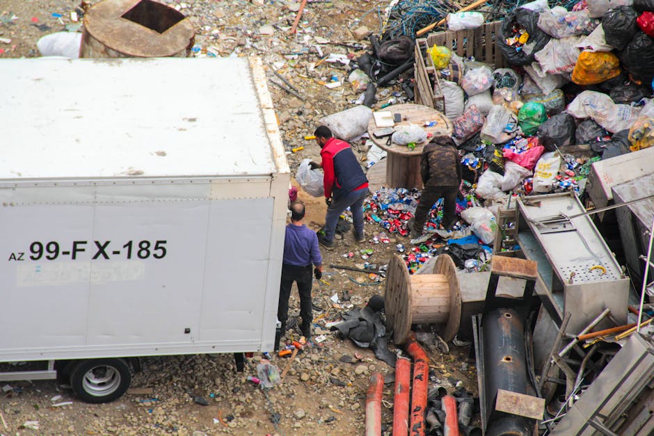 A large green waste collection bin filled with various types of cardboard boxes, packaging materials, and other mixed rubbish, located on a city street with a building and trees in the background. The